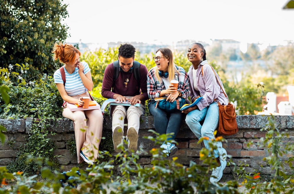 A young group of friends sitting on a stone ledge outside, laughing and smiling as they study together.