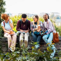 A young group of friends sitting on a stone ledge outside, laughing and smiling as they study together.