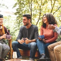 A group of friends sitting on an outdoor patio with mugs with a backdrop of trees