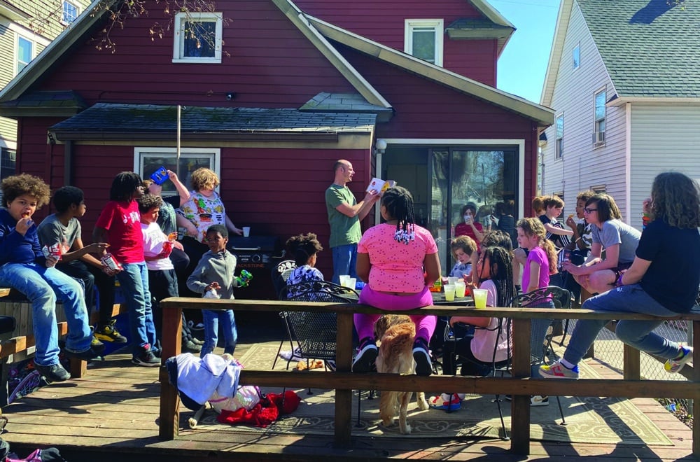 A diverse group of kids sit on a sunny patio attached to a red house as a man reads from the Bible.