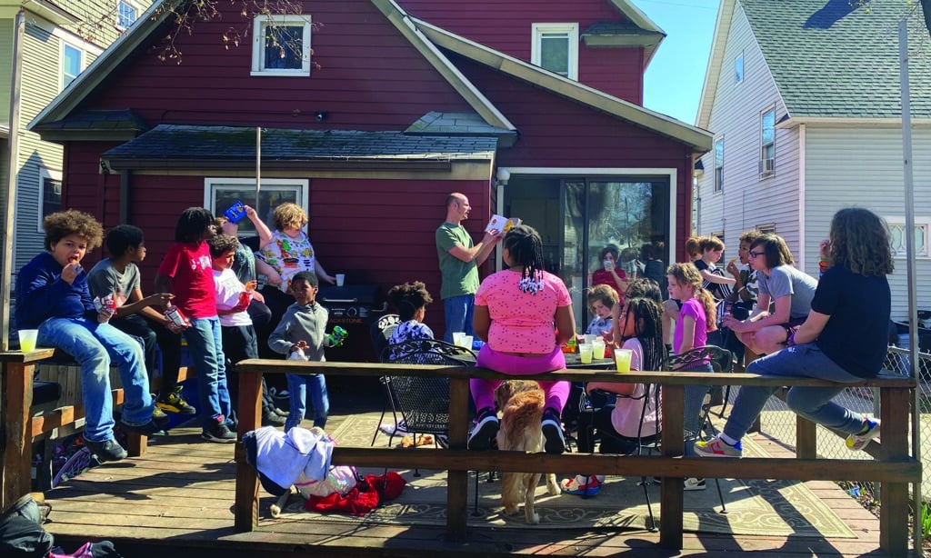 A diverse group of kids sit on a sunny patio attached to a red house as a man reads from the Bible.