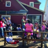 A diverse group of kids sit on a sunny patio attached to a red house as a man reads from the Bible.