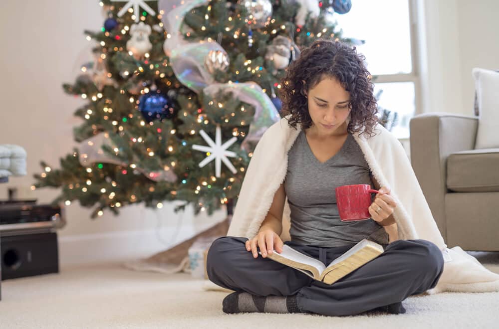 A woman sits on the floor near her Christmas tree, wrapped in a blanket reading Isaiah 9:6 in her Bible.