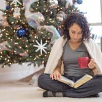 A woman sits on the floor near her Christmas tree, wrapped in a blanket reading Isaiah 9:6 in her Bible.