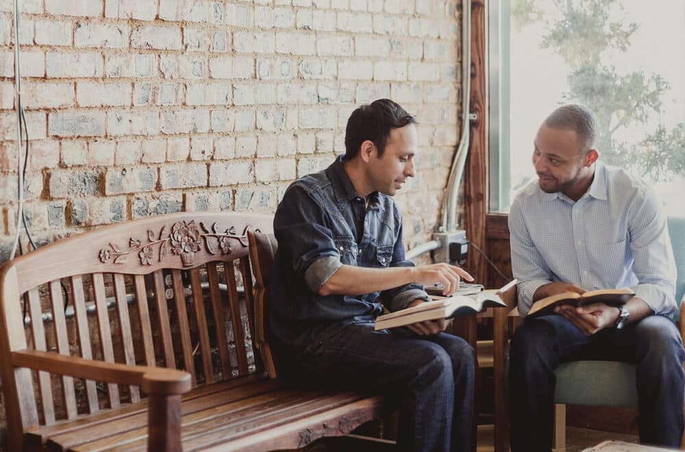 Two men sit on a bench, reading the Bible together.