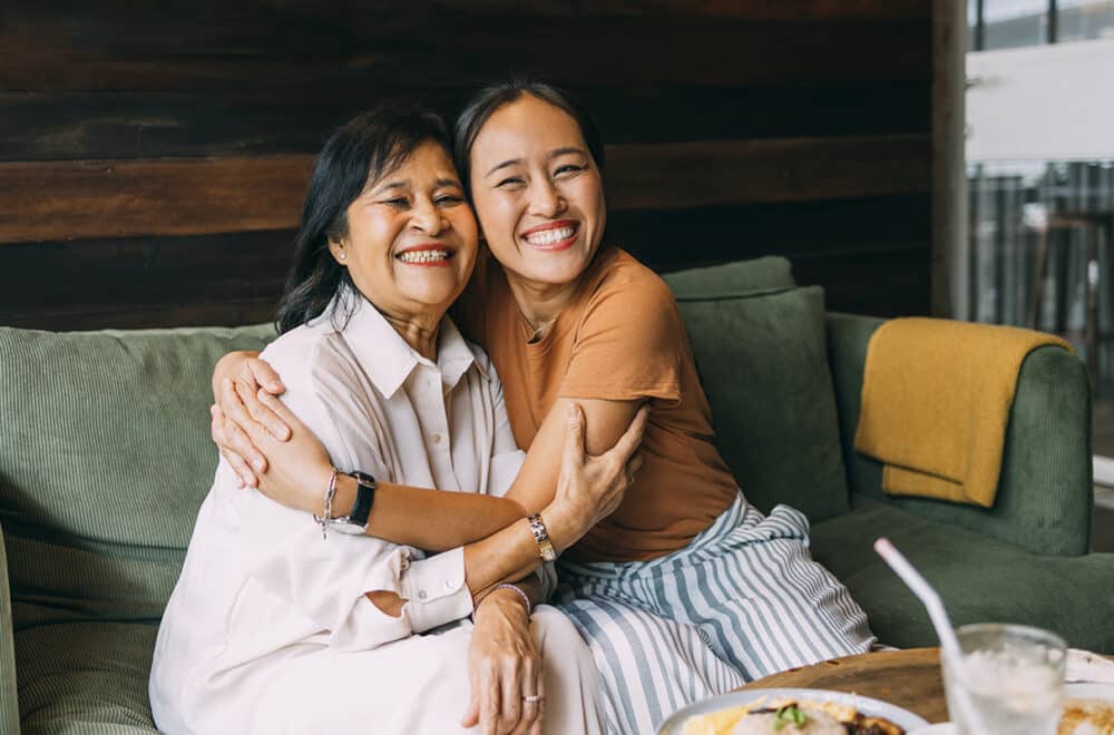 Two Thai women sit in coffee shop on a green couch, embracing one another.