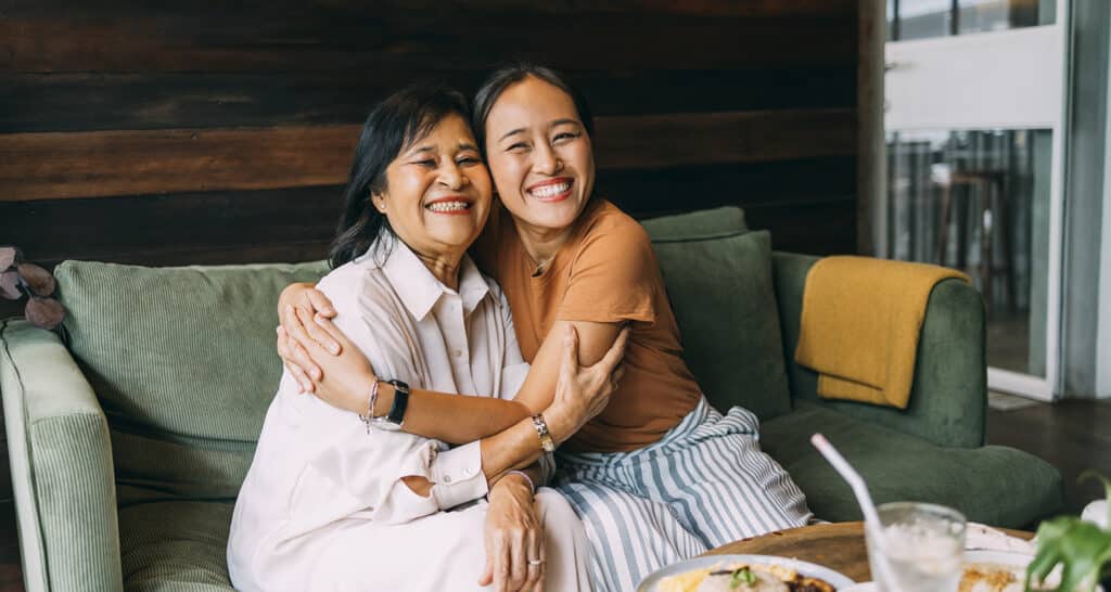 Two Thai women sit in coffee shop on a green couch, embracing one another.