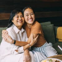 Two Thai women sit in coffee shop on a green couch, embracing one another.