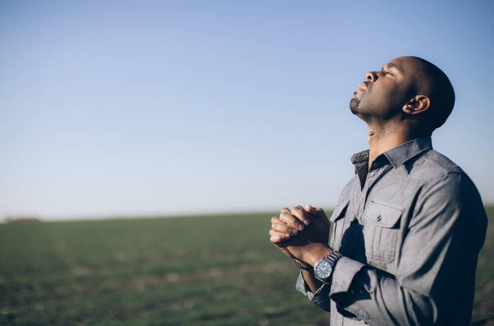 A man praying in an open field with his hands folded and head pointed toward the sky.