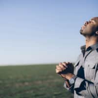 A man praying in an open field with his hands folded and head pointed toward the sky.