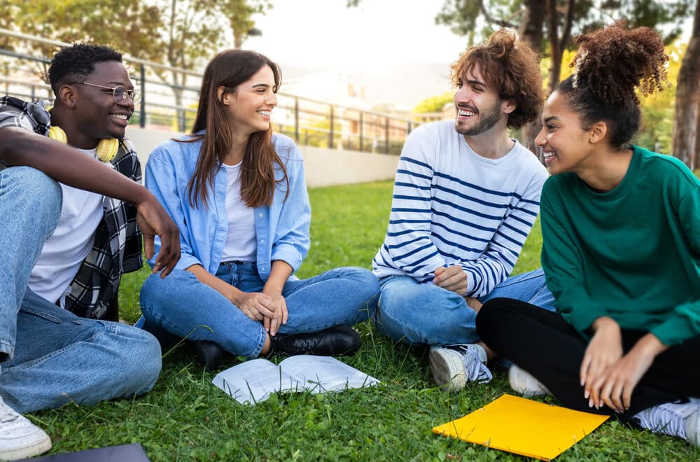 A group of four friends sit in the grass smiling and reading their Bible at a park.