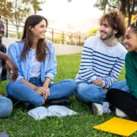 A group of four friends sit in the grass smiling and reading their Bible at a park.